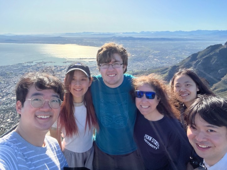 group of six people taking a selfie on top of a mountain with the ocean in the background