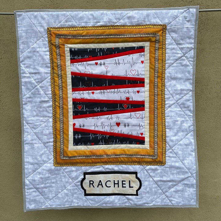 hanging quilt with a gold frame that shows health-themed fabric and the name plate of Rachel underneath