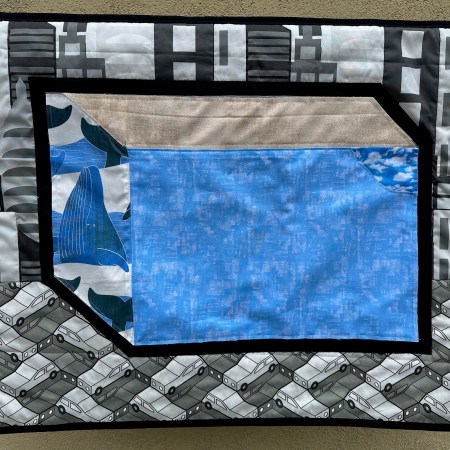 Hanging quilt of blue building with a whale on the side. Background is city and parked cars.